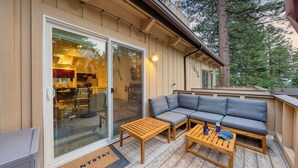 Un espacio al aire libre acogedor con un sofá y dos mesas de madera, una puerta de cristal que da a la cocina, y una terraza de madera.