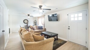 Een woonkamer met een beige bank, een bijzettafeltje, een plafondventilator en een flatscreen-televisie aan de muur.