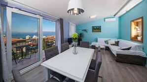 Ein modernes Wohnzimmer mit einer großen Schiebeglaswand, die auf einen Balkon führt, einem weißen Esstisch, grauen Sofas und Blick auf das Meer.