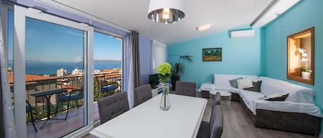 A modern living room with a large sliding glass door leading to a balcony, a white dining table, grey sofas, and a view of the sea.