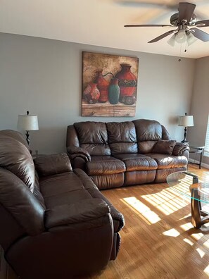 Ein Wohnzimmer mit braunem Ledersofa, einem Glastisch und einem Deckenventilator.