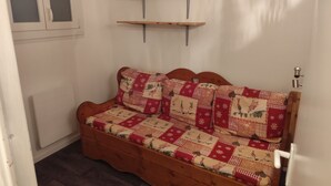 Ett mysigt rum med en soffa som kan fällas ut till en säng, två hyllor i trä, ett element och en dörr med ett handtag.