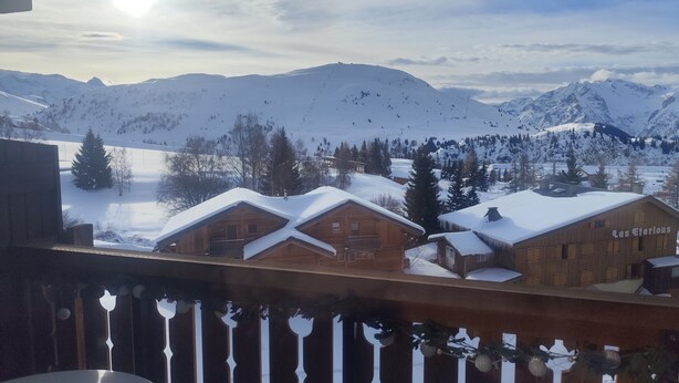 En stuga av trä med ett snötäckt tak, omgiven av berg och en klar himmel.
