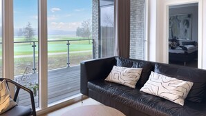 A modern living room with a leather sofa, a view of a golf course, and a balcony with a glass door. - Lübker Golf og Spa Resort (Nimtofte)
