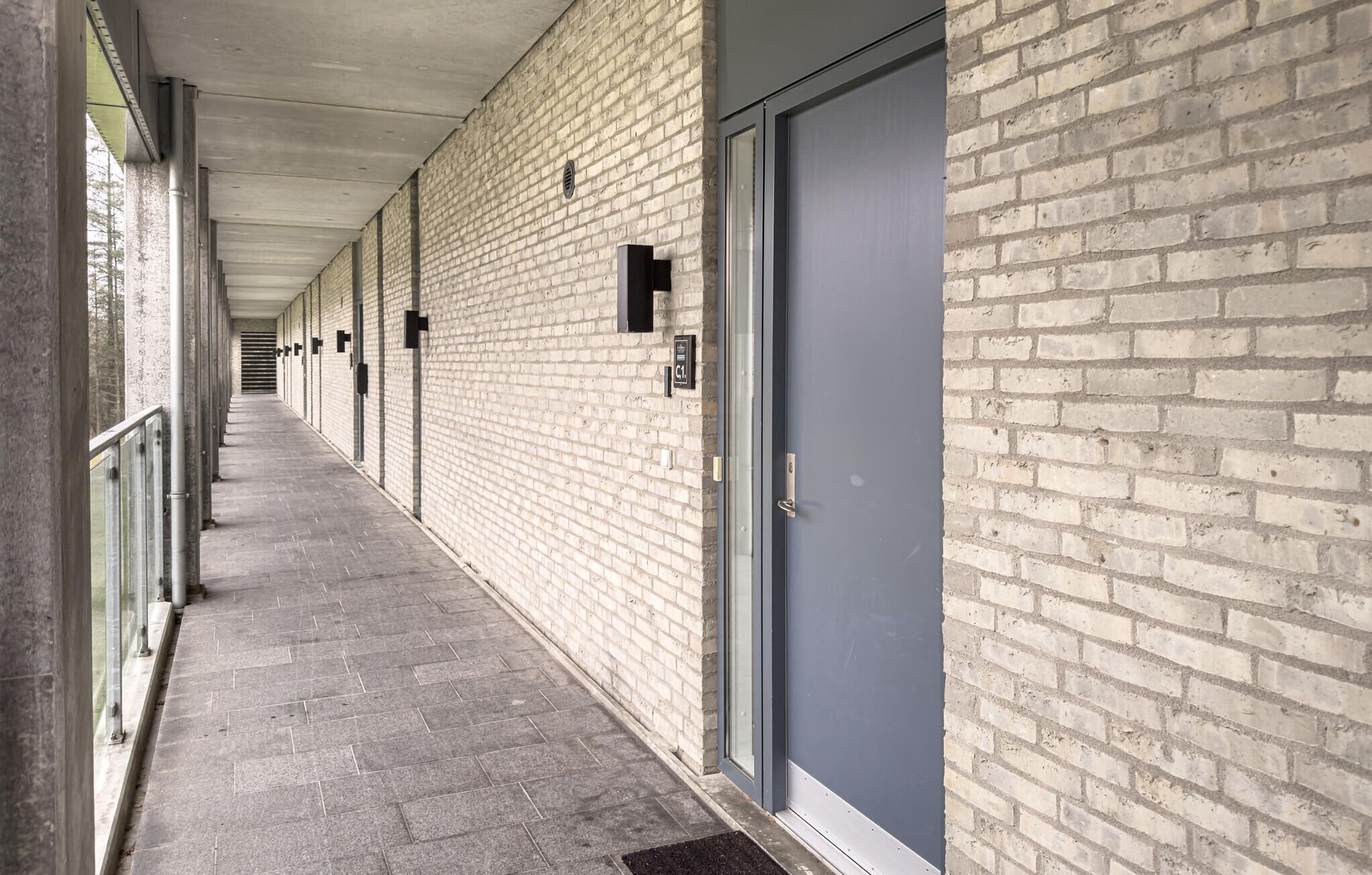 A corridor with a brick wall, a grey door, and a metal railing.