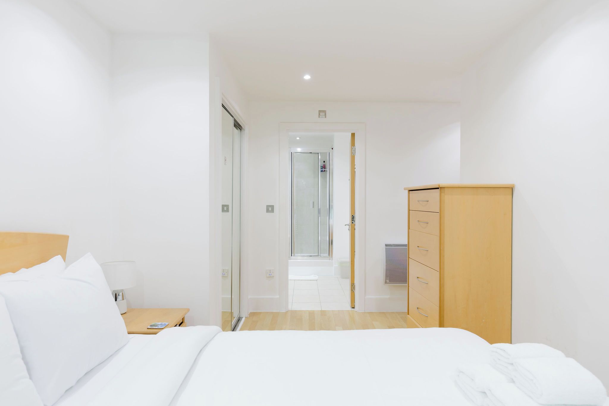 A hotel room with a bed, a wooden wardrobe, a nightstand, and a bathroom visible through a glass door.