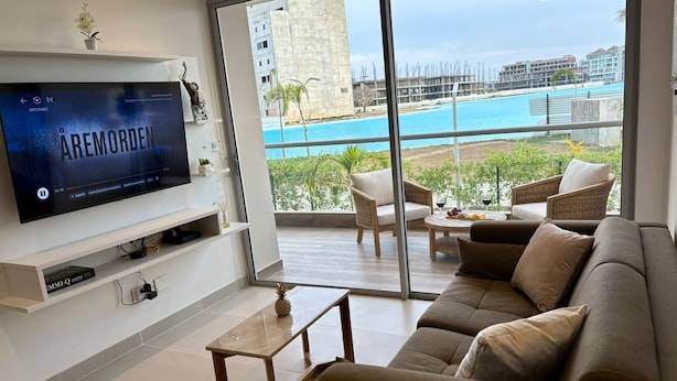 Ein modernes Wohnzimmer mit großem Fernseher, bequemen Sitzgelegenheiten und einem Balkon mit Blick auf einen Pool und Gebäude.