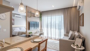 A modern living room with a sofa, a dining area with a table and chairs, a TV, and a large window with curtains. - Cozy Studio in Cambuí (Campinas)