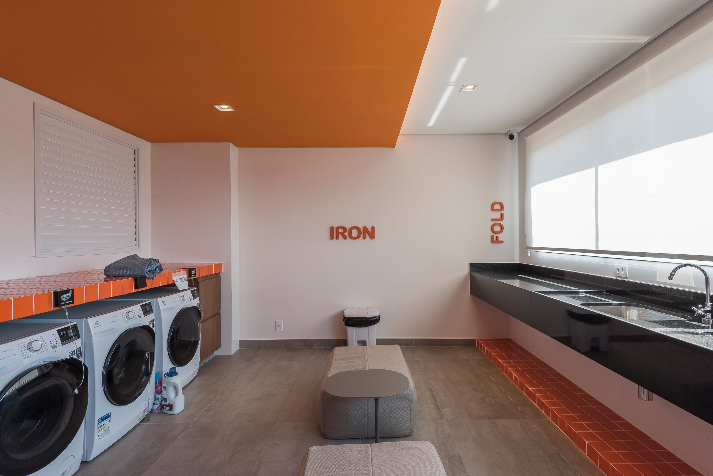 A modern laundry room with two front-loading washing machines, a small stool, a bench, and a large window providing natural light.