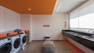 A modern laundry room with two front-loading washing machines, a small stool, a bench, and a large window providing natural light.