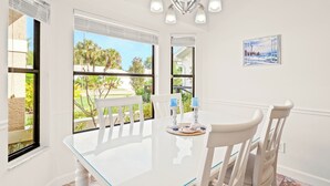Un comedor con una mesa y sillas blancas, ventanas amplias con persianas y una araña.