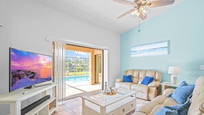 A modern living room with a flat-screen TV, a glass-top coffee table, a beige sofa, and a ceiling fan. - Peaceful Lakeview 3BR. Heated Pool Retreat.  (Naples)
