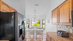 A kitchen with wooden cabinets, a black refrigerator, a toaster, and a dining area with a table and chairs. - Peaceful Lakeview 3BR. Heated Pool Retreat.  (Naples)