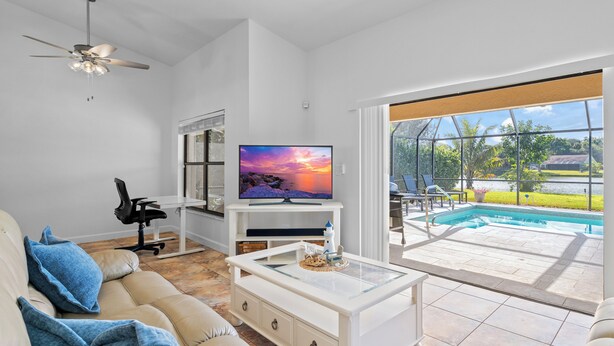 A living room with a TV, sofa, and coffee table, a ceiling fan, and a view of a swimming pool. - Peaceful Lakeview 3BR. Heated Pool Retreat.  (Naples)