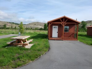A small wooden cabin with a door and windows, a picnic table, and a paved path leading to it.