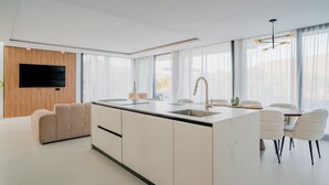 Una cocina moderna con una isla central, un comedor con sillas blancas y un televisor de pantalla plana montado en un panel de madera.