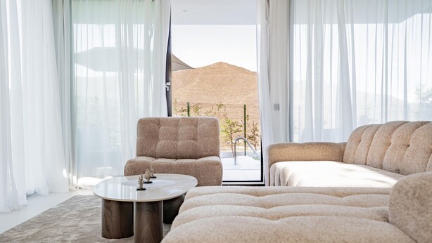 Una sala de estar moderna con sofá, sillón y mesa de centro, ventanas amplias con cortinas translúcidas y vistas a una colina.