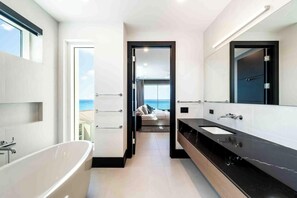 Un baño moderno con bañera, un espejo grande y vistas al mar a través de una puerta abierta.