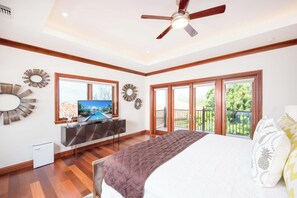 A bedroom with a bed, a TV, a ceiling fan, and large windows with a view of greenery. - Casa Luna (George Town)