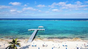 Une plage à l’eau turquoise et limpide, une jetée menant à un kiosque, et des chaises longues sur le sable.