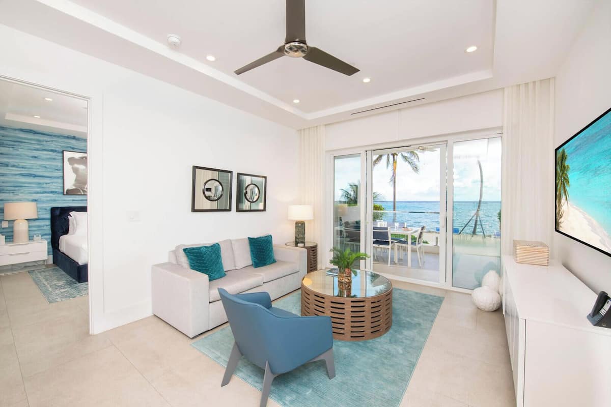 A modern living room with a sofa, armchair, and coffee table, a TV, and a balcony with a view of the ocean.