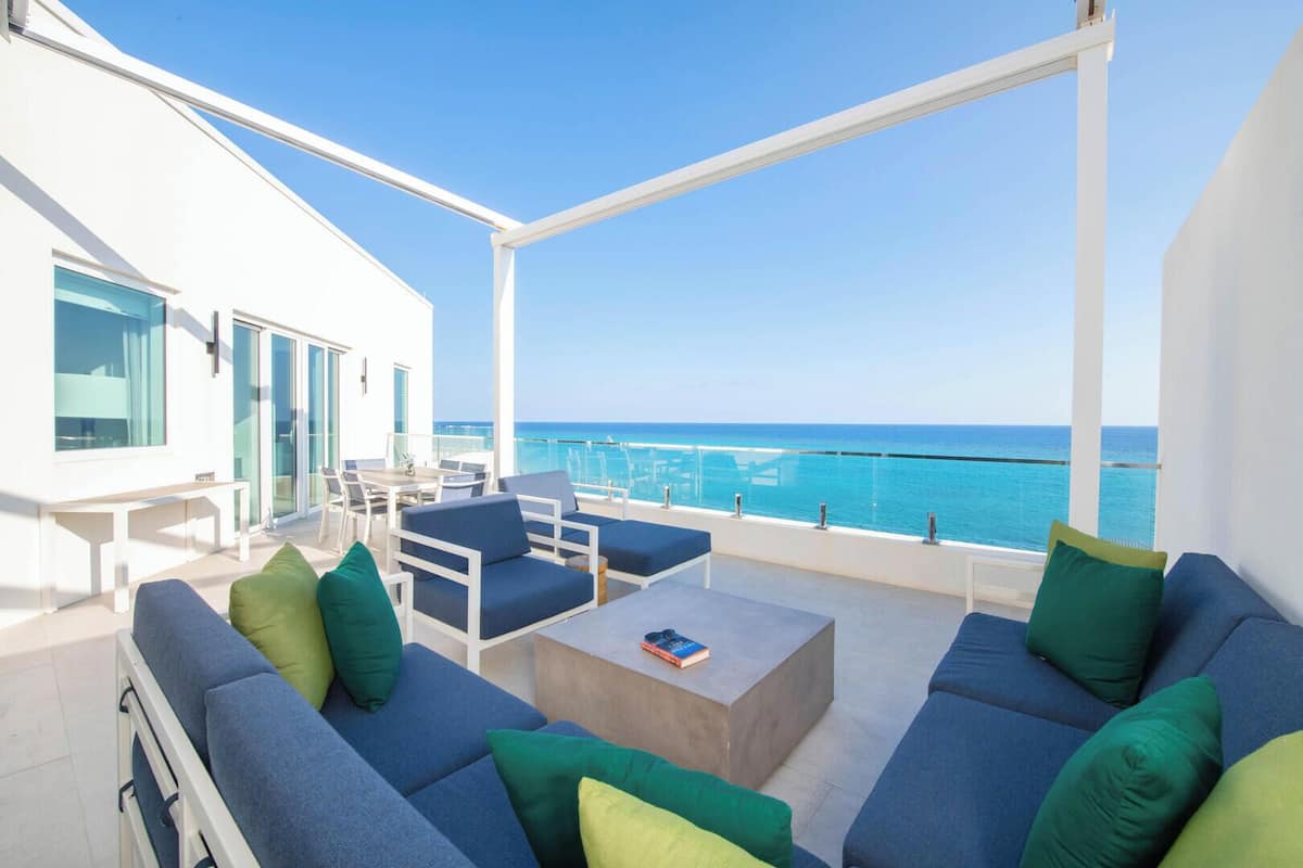 A modern outdoor seating area with blue sofas and green cushions, overlooking a clear blue sea.