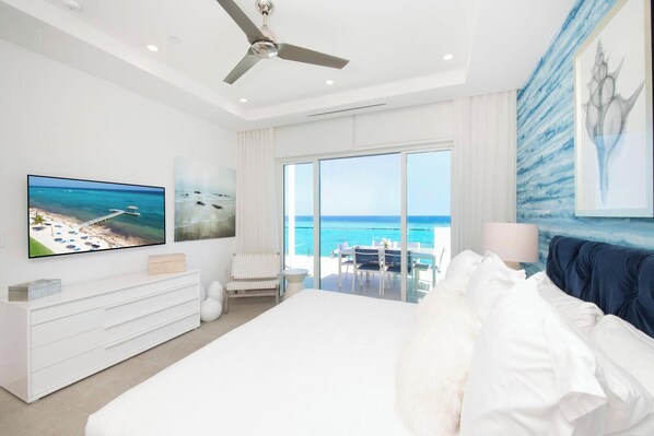 Een slaapkamer met een groot bed, een televisie, een plafondventilator en uitzicht op de oceaan door grote ramen.