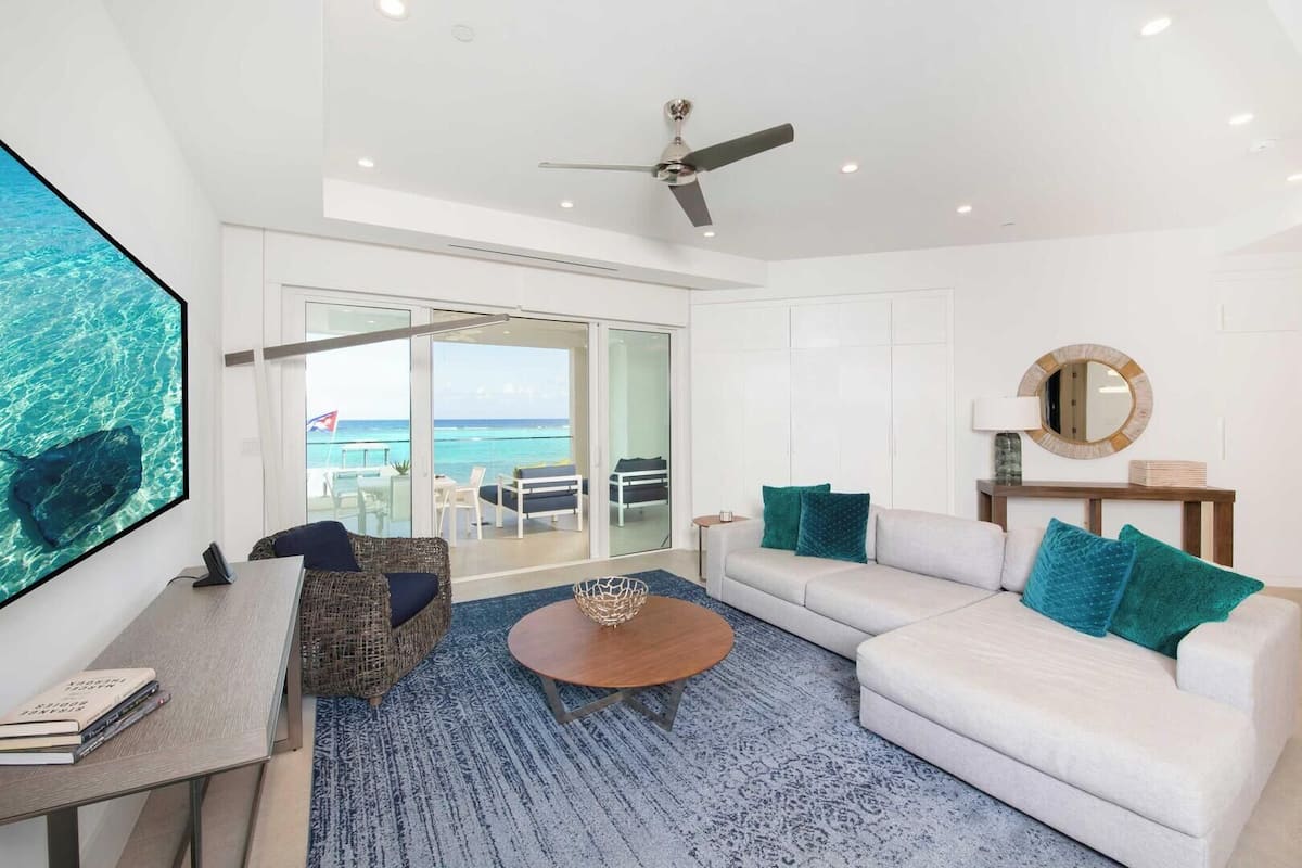 A modern living room with a large TV, a ceiling fan, a sofa, a coffee table, and a view of the ocean through large glass doors.