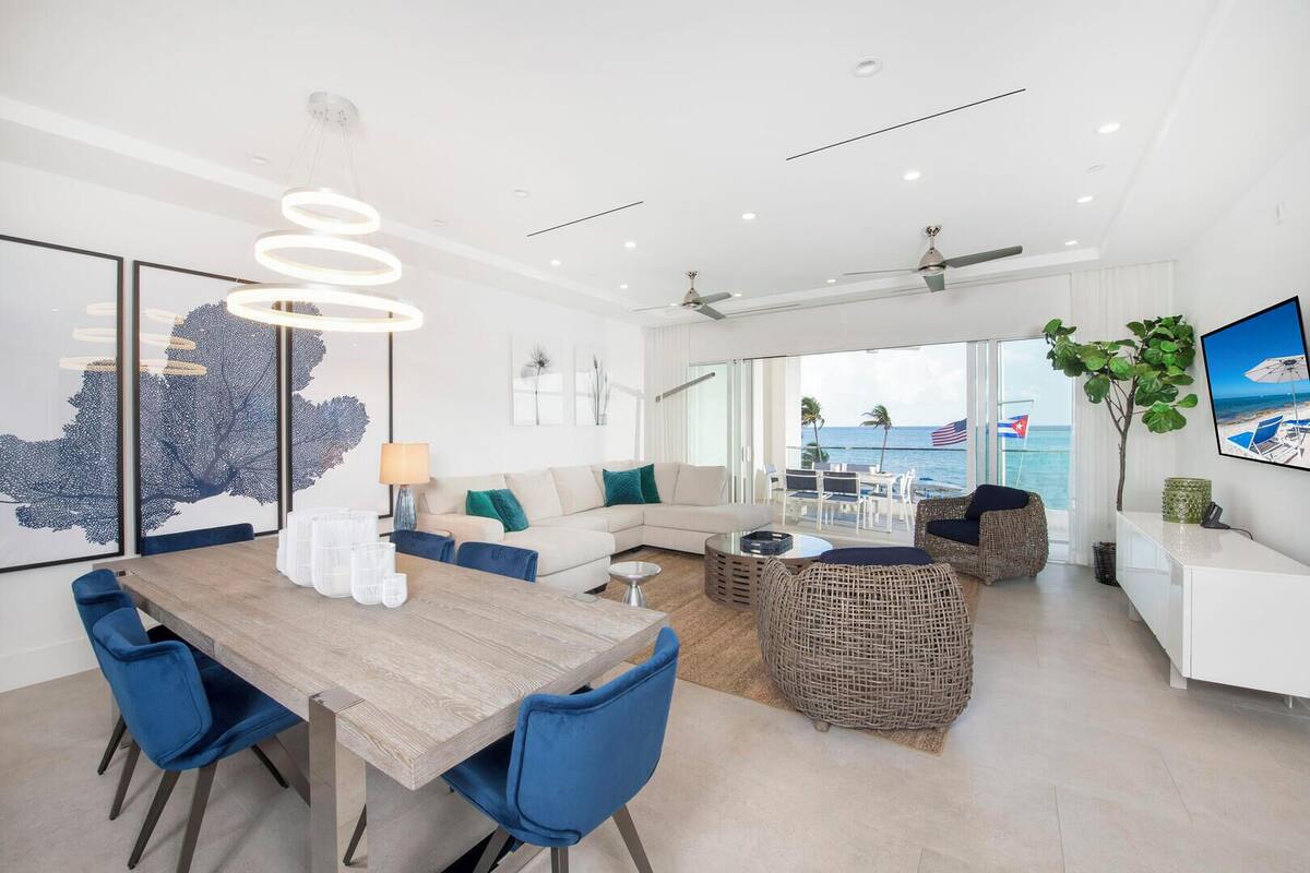 A modern living room with a wooden dining table, blue chairs, a sofa, and a balcony with a view of the ocean.