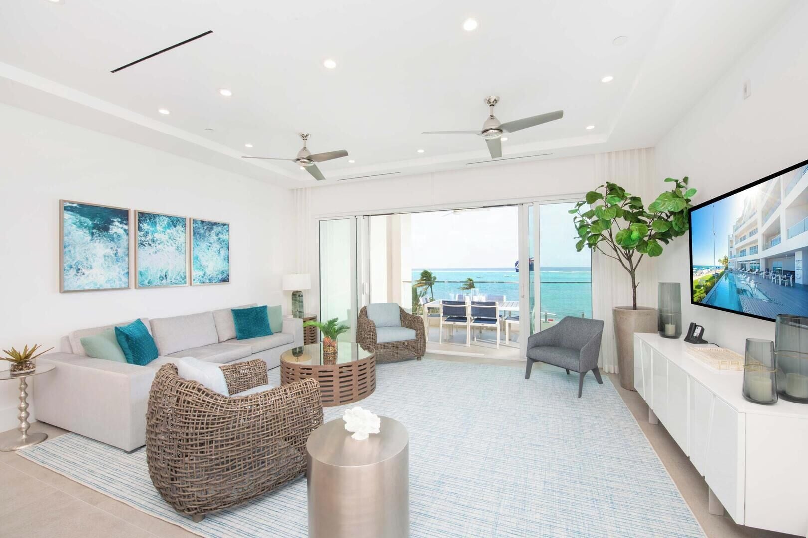 A modern living room with a sofa, wicker chairs, a coffee table, a TV, and a balcony with a view of the ocean.