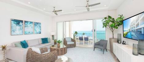 A modern living room with a sofa, wicker chairs, a coffee table, a TV, and a balcony with a view of the ocean.