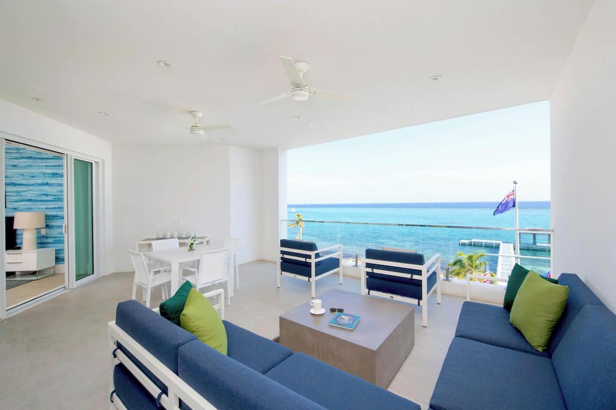 A modern living room with a balcony overlooking the sea, featuring a sofa set, a dining table, and ceiling fans.