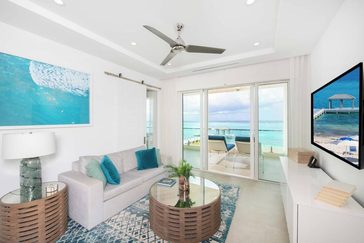 A modern living room with a sofa, coffee table, and TV, overlooking a balcony with a view of the ocean.
