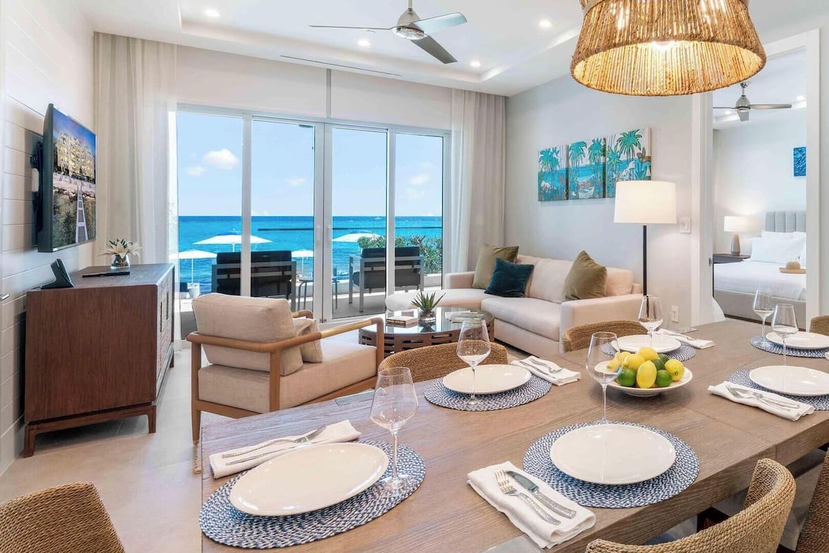A modern hotel room with a dining area, a sofa, a TV, and a view of the ocean.