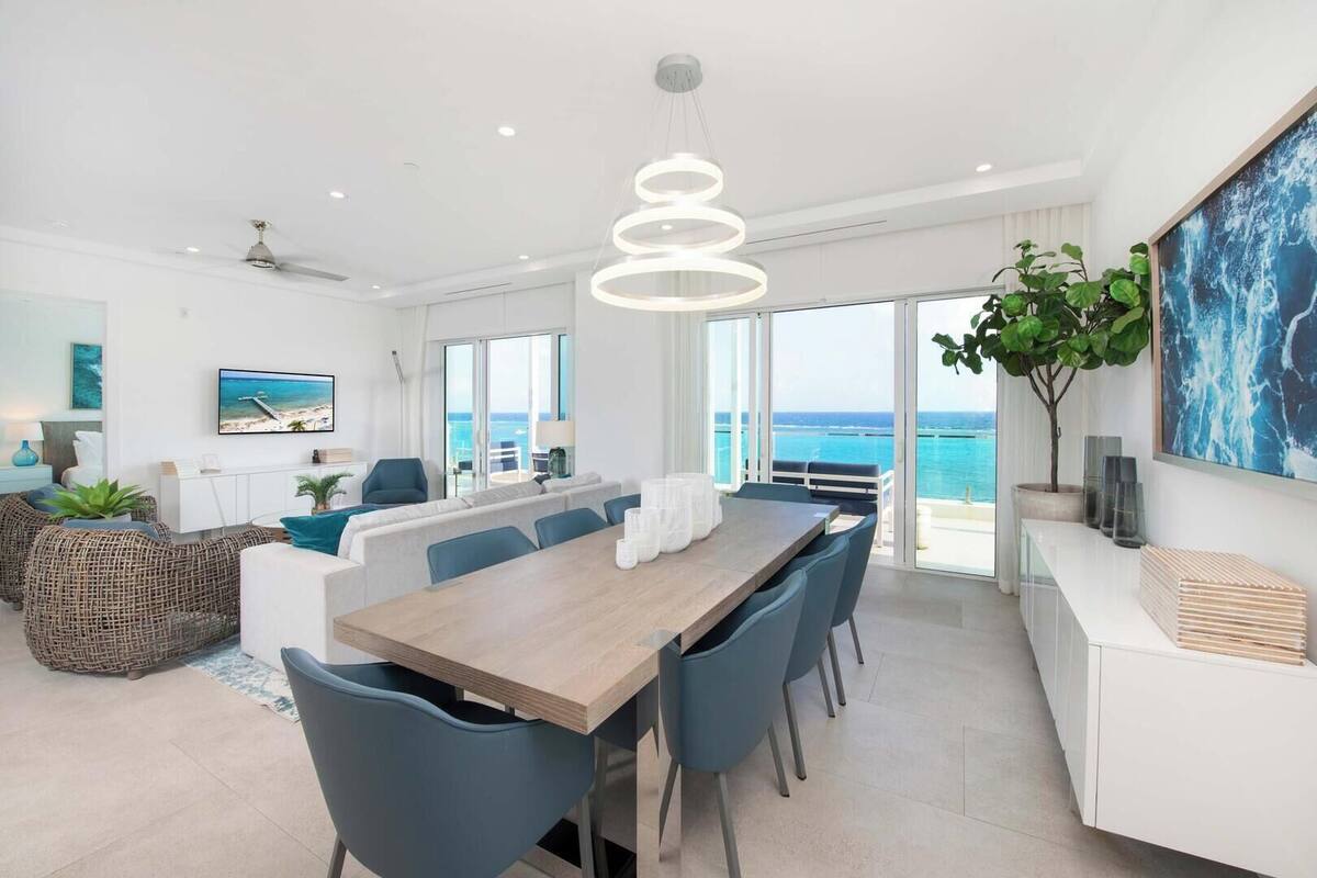 A modern dining area with a wooden table and blue chairs, large windows with a view of the sea, and a hanging light fixture.