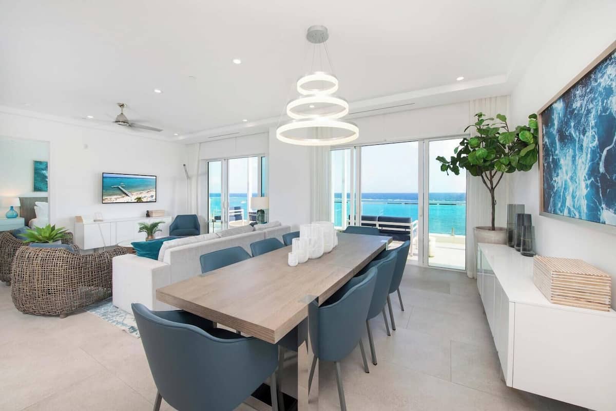Un comedor moderno con una mesa de madera y sillas azules, ventanas amplias con vista al mar y una lámpara colgante.