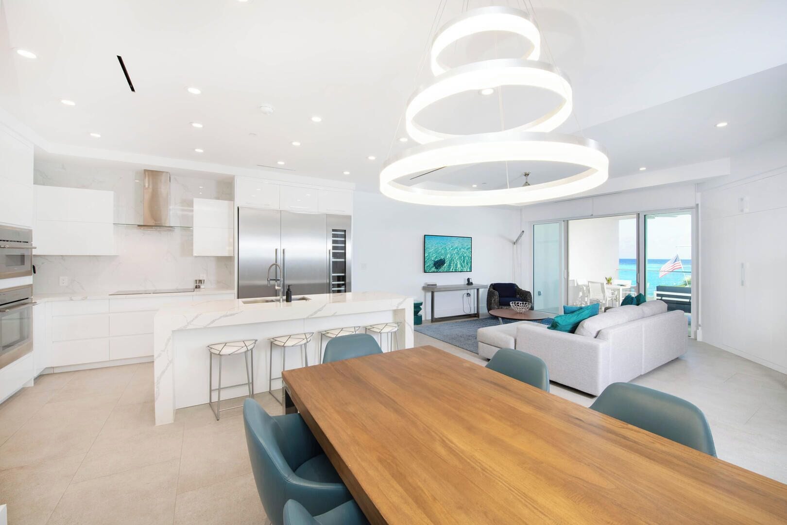 A modern kitchen with a central island, a dining area with a wooden table and chairs, and a living area with a sofa and a TV.