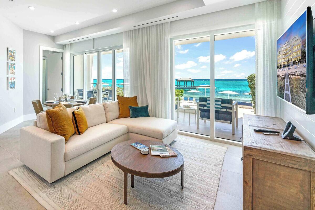 A modern living room with a large sofa, a coffee table, and a TV. There is a balcony with a view of the ocean.