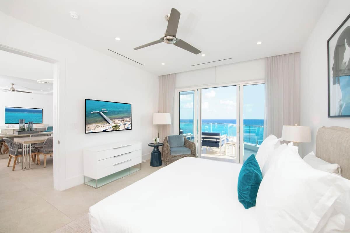 A modern hotel room with a balcony, a TV, a desk, and a bed with white and teal pillows.