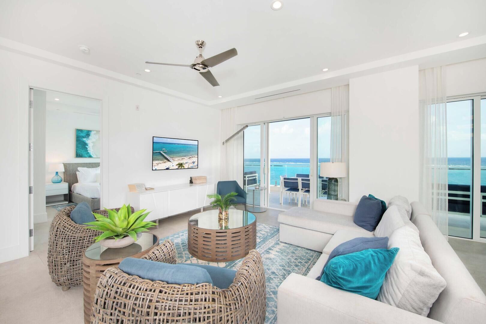A modern living room with a ceiling fan, a large window with a view of the ocean, wicker furniture, and a white sofa with blue cushions.