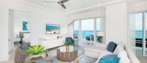A modern living room with a ceiling fan, a large window with a view of the ocean, wicker furniture, and a white sofa with blue cushions.