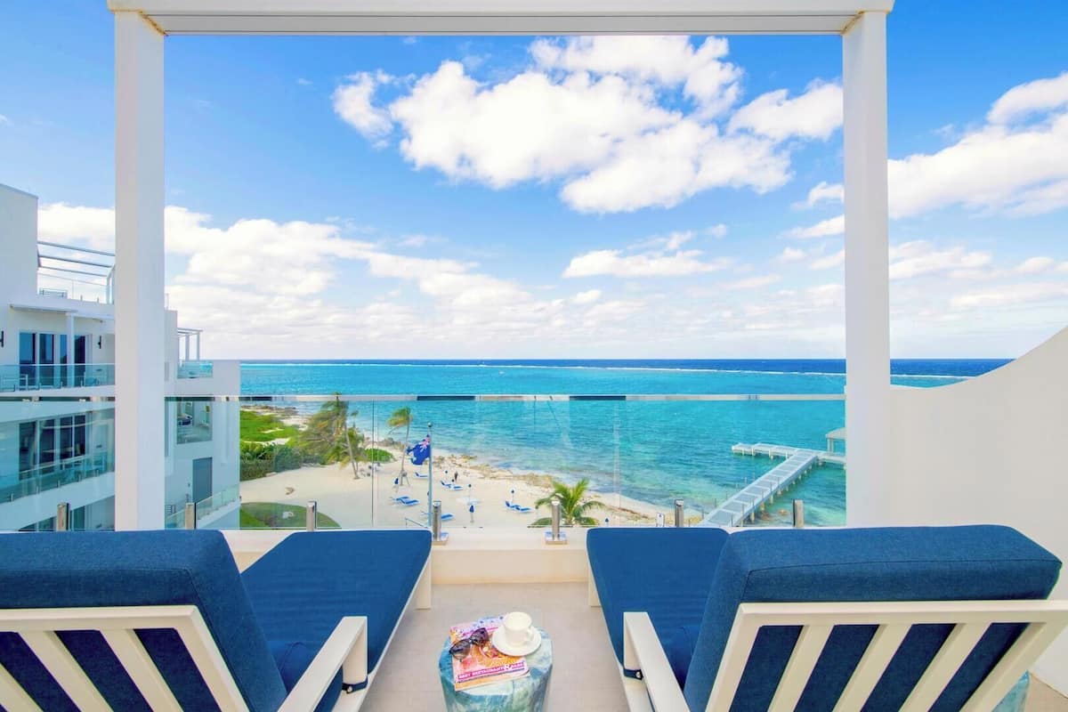 A balcony with two blue chairs, a small table with a magazine and a cup, and a view of the ocean and beach.
