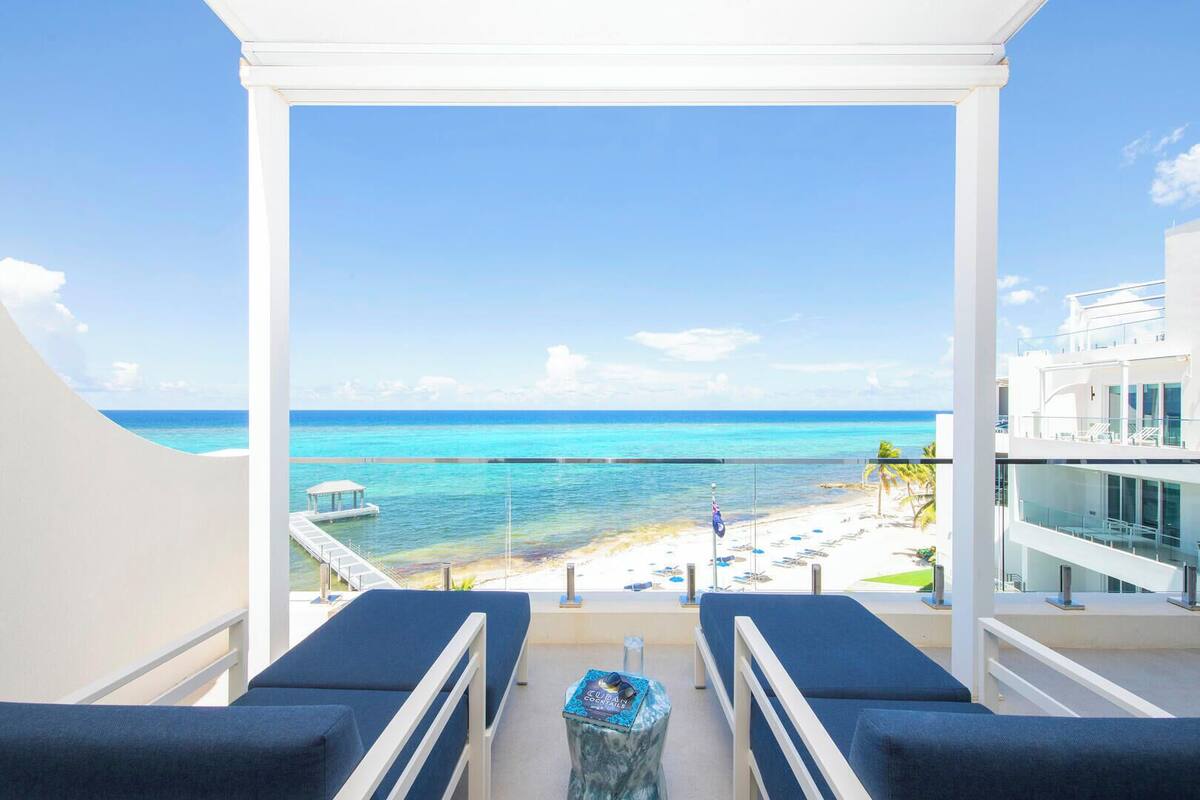 A balcony with a view of the beach and ocean, a lounge area with blue sofas, and a small table with a drink.