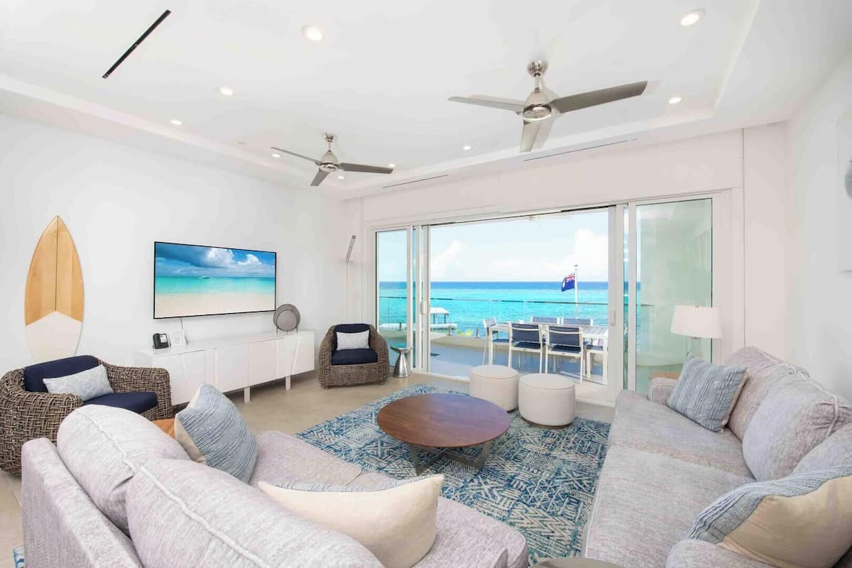 A spacious living room with a large TV, comfortable seating, ceiling fans, and a balcony with a view of the ocean.