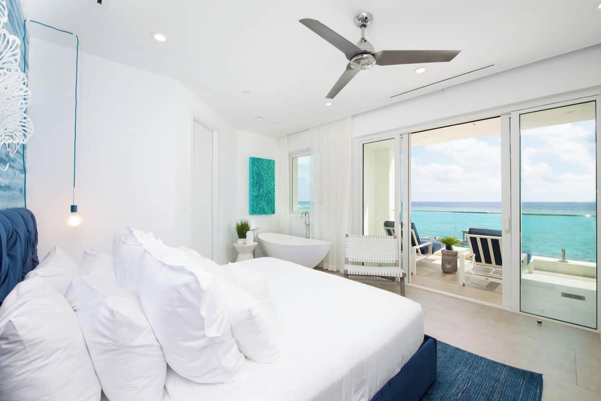 A modern bedroom with a large bed, a ceiling fan, a bathtub, and a balcony with a view of the ocean.