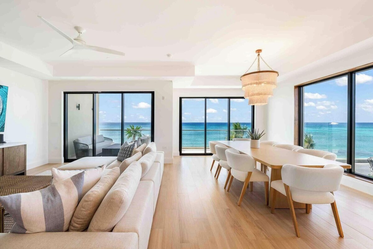 Ein modernes Wohnzimmer mit Sofa, Esstisch und Stühlen, großen Fenstern mit Meerblick und einem Deckenventilator.