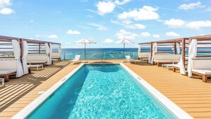 Un espace aménagé au bord de la piscine, avec des chaises longues et des parasols, offrant une vue dégagée sur l’océan, ainsi qu’une terrasse en bois.