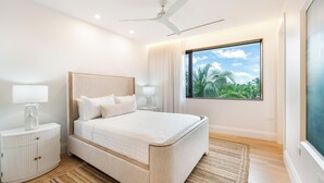 Une chambre moderne avec un grand lit, un ventilateur de plafond, une table de chevet et une fenêtre donnant sur des palmiers.
