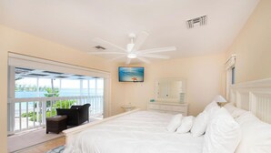 A bedroom with a large bed, a ceiling fan, a painting, a dresser, and a view of the ocean through a sliding door. - Pools of the Kai (North Side)