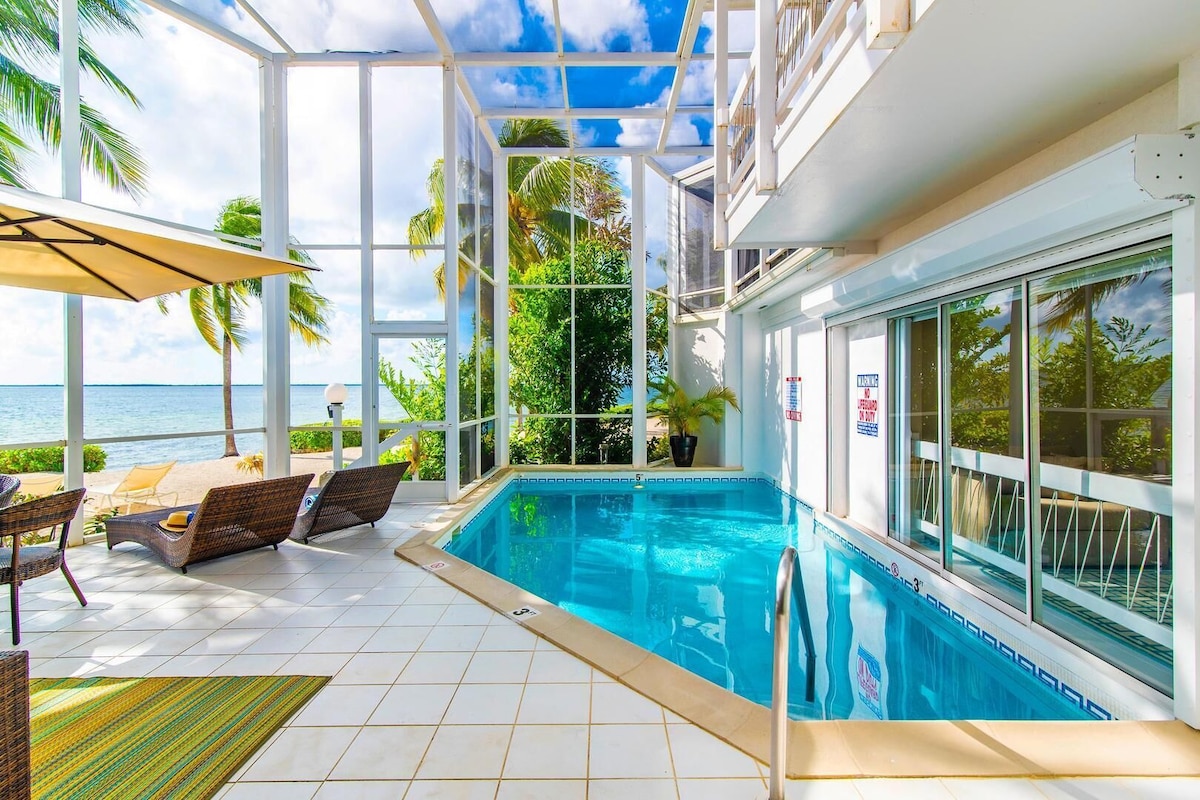 A pool area with a view of the ocean, a covered patio with chairs, and a glass-enclosed structure.